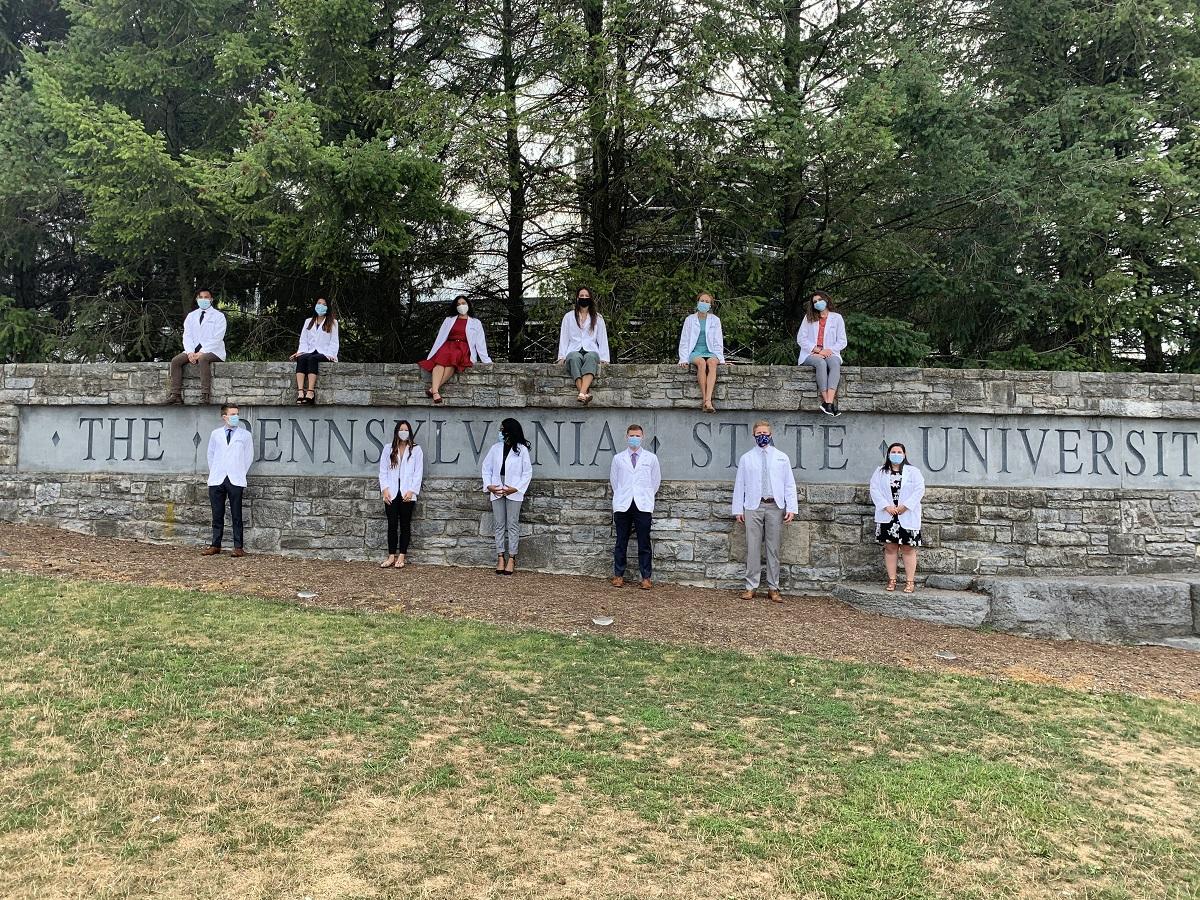 Medical school students wearing white physician coats stand and sit around a gray, stone wall with the words “Penn State University” carved into it. A row of trees stand in the background.