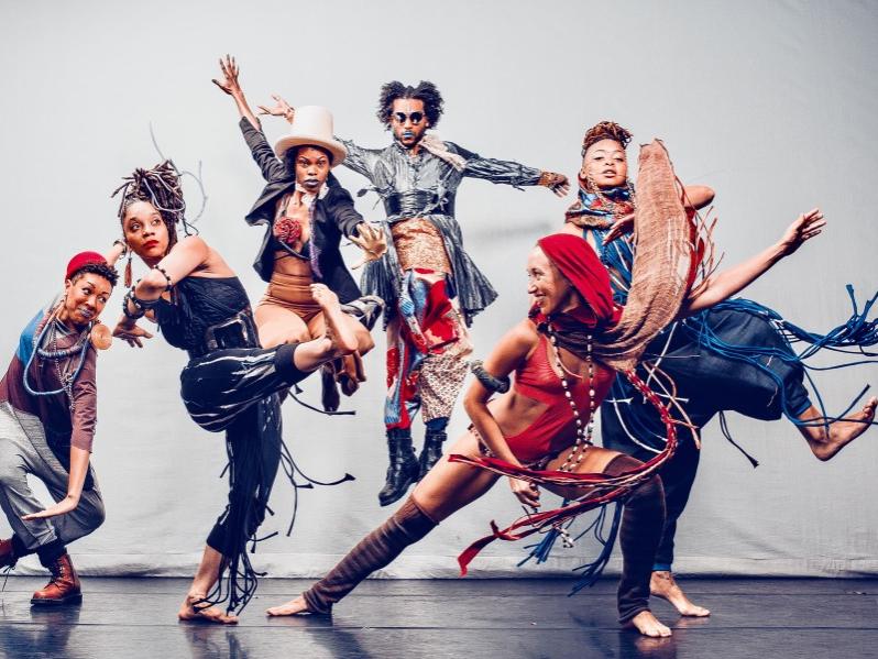 Six dancers—each sporting a braided or otherwise interesting hairstyle and baggy and tassled costumes—are shown in suspended jumps and kicks.
