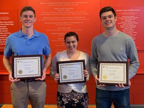 Three students, two male, one female, each holding framed Undergraduate Exhibition award in front of orange background. 