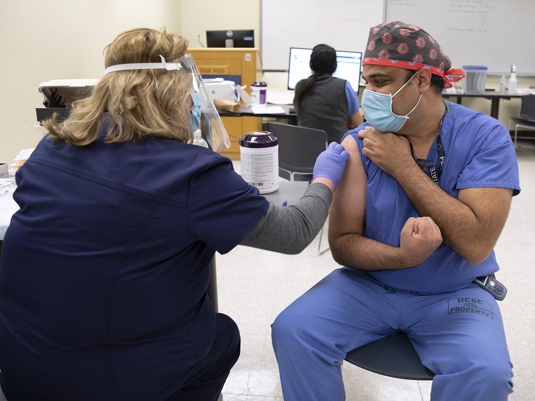 A man in scrubs, a head covering and a surgical masks, clenches his fist, while a woman in personal protective equipment prepares to give him a shot.