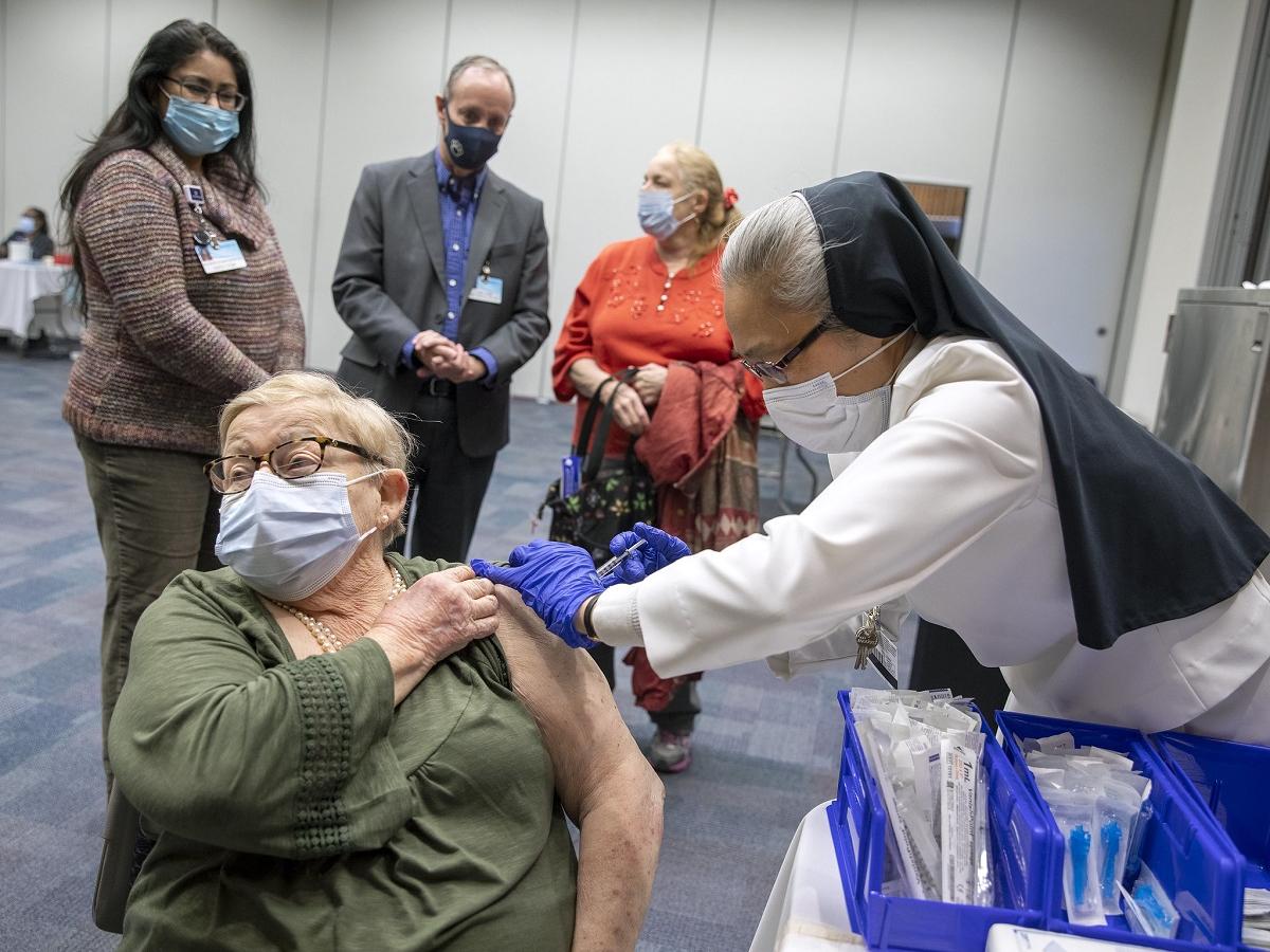 A woman in a mask looks away as another woman in hospital scrubs, rubber gloves and a nun’s habit gives her in an injection into her shoulder.