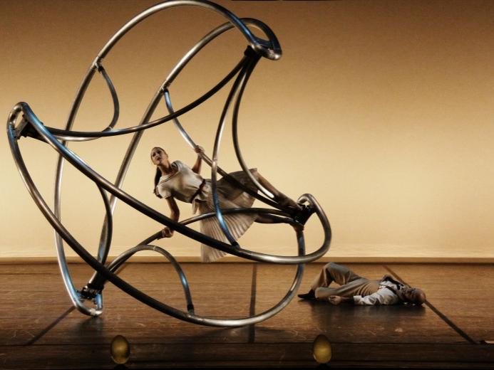 An acrobat is positioned on the Cyr Wheel while another lies on the ground nearby.