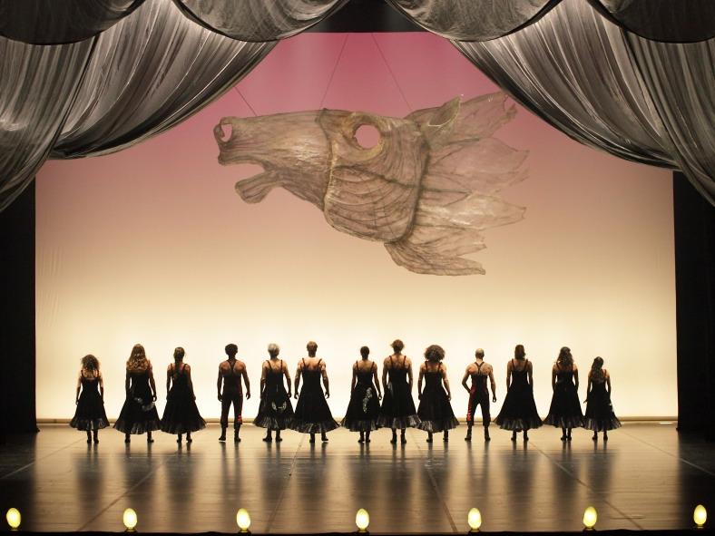 Members of Compagnia Finzi Pasca stand in a line with their backs to the audience while a large sculpture of a horse head is suspended above the acrobats in a scene from the production of “La Verità.”