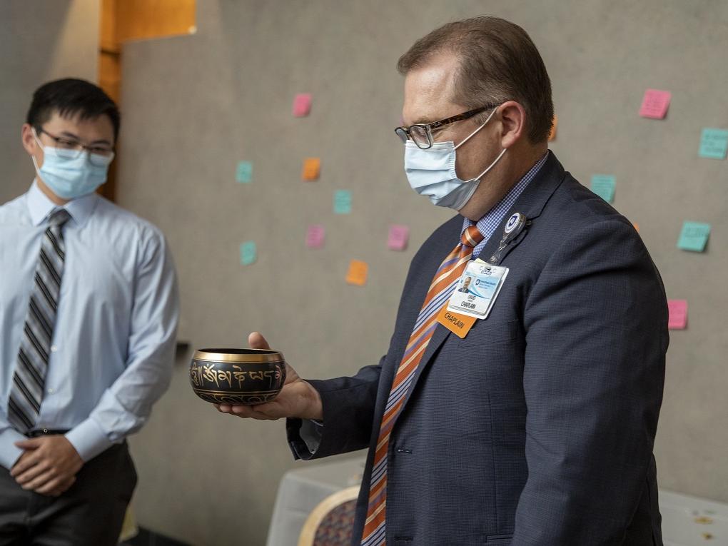 David Simmons stands holding a bowl in one hand. Behind him, another man in a mask wearing a tie and shirtsleeves, watches. They stand in front a wall covered with notes.