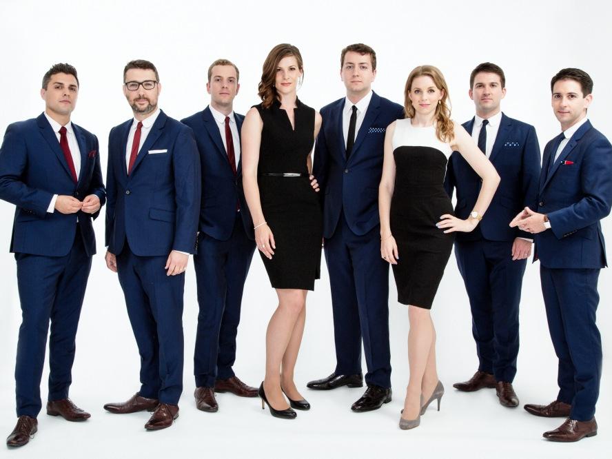 The eight singers of vocal group Voces8 wear casual suits and dresses and stand shoulder to shoulder.