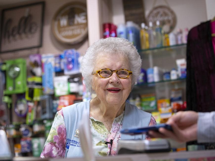 Arlene Faul, 95, from Hershey, smiles behind the counter of the Hershey Medical Center gift shop. She is wearing a vest, shirt, necklace and glasses. A man’s hand holds a phone out to her. Behind her are shelves of toiletries and other items.