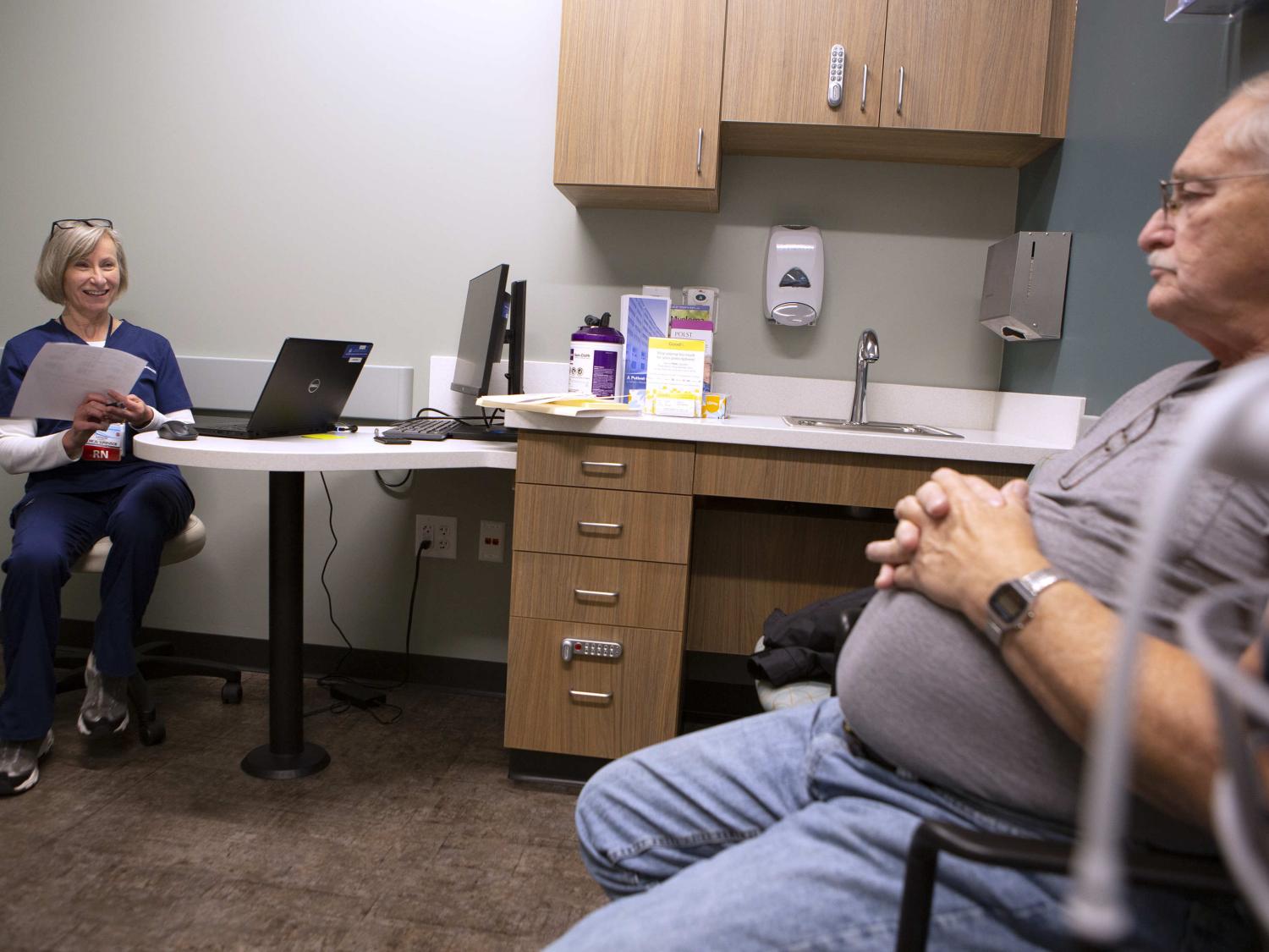 Kat Schoenknecht sits at a desk in a doctor’s office with a laptop opened. She holds a sheet of paper and smiles at a man seated across from, who sits with his hands folded on his stomach.