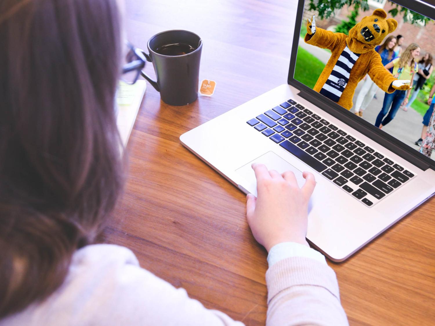 A student sits at a desk with a mug of hot tea, a cell phone, and a computer with Penn State's Nittany Lion mascot welcoming her on the screen.