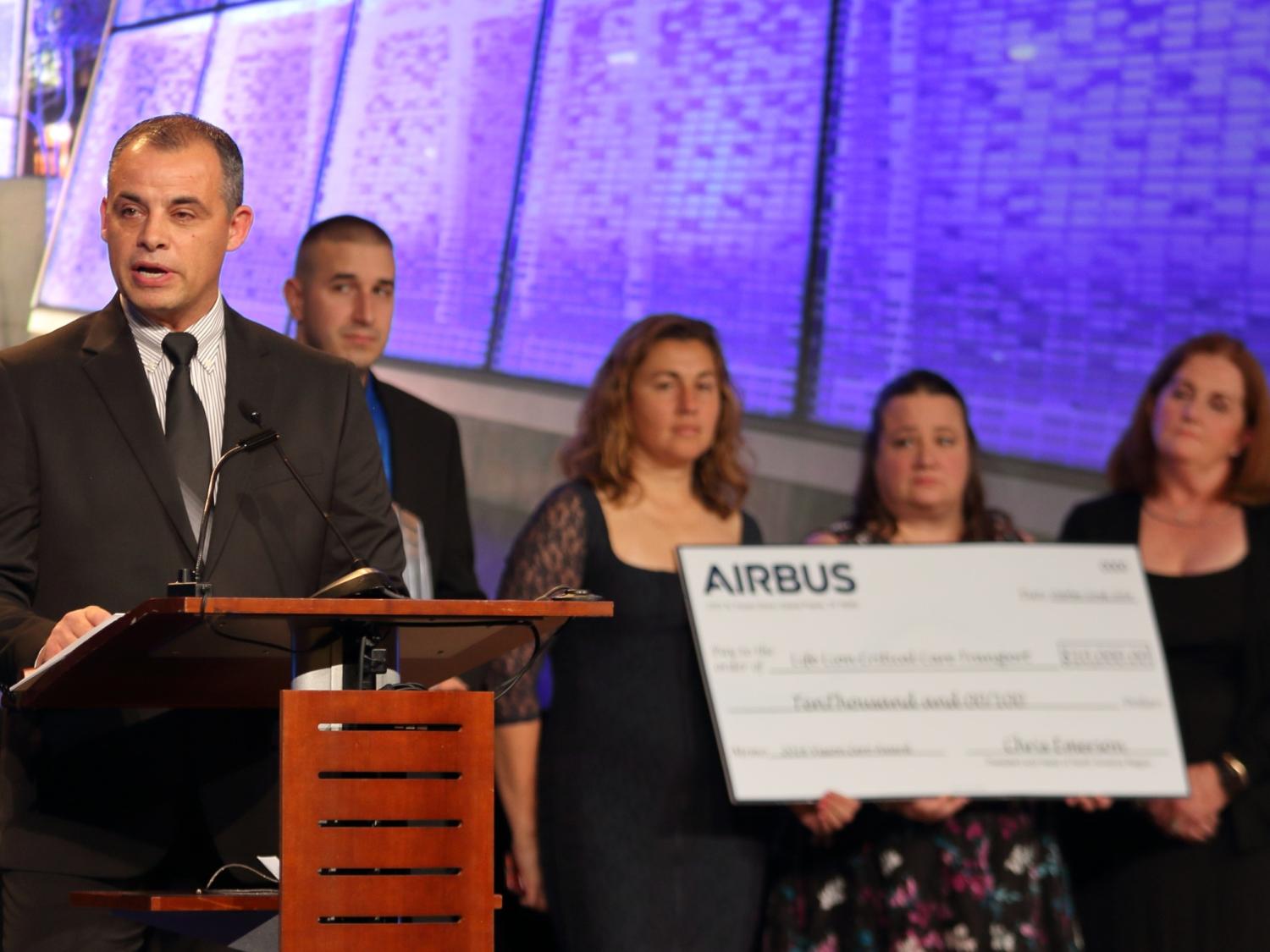A man in a suit stands at a lectern speaking. Four people stand behind him holding an oversized cardboard check.