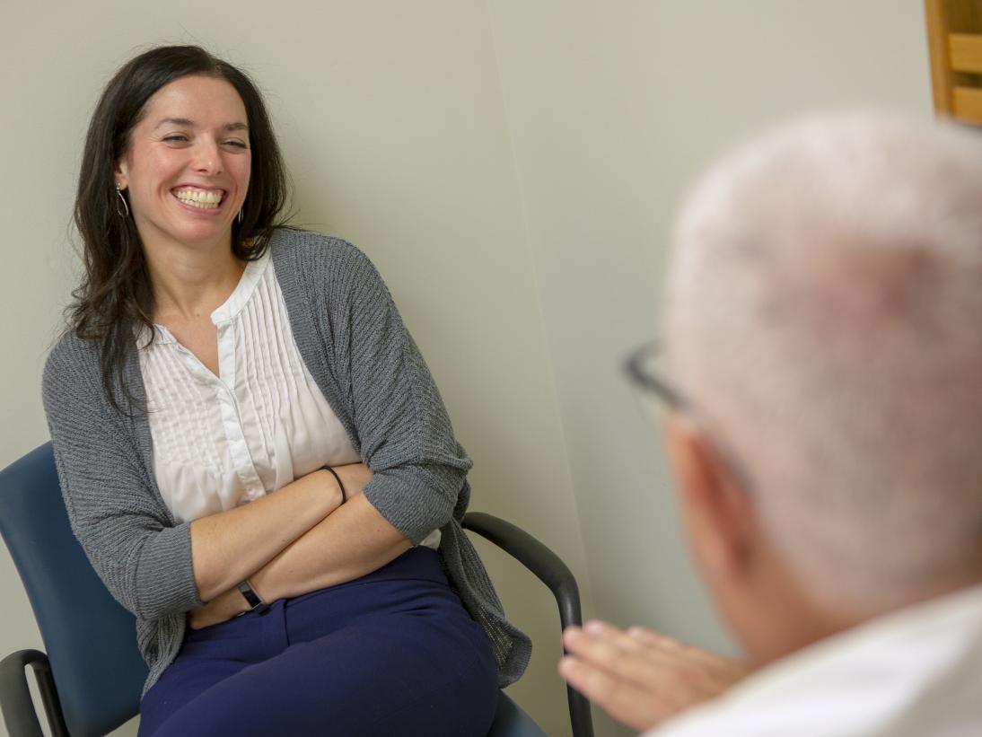 Princess Young smiles as she listens to Dr. Gerald Harkins during an endometriosis preoperative appointment. She has long hair and is wearing a sweater, top and casual pants. He is seen from behind. He has white hair and is wearing glasses and a lab coat.