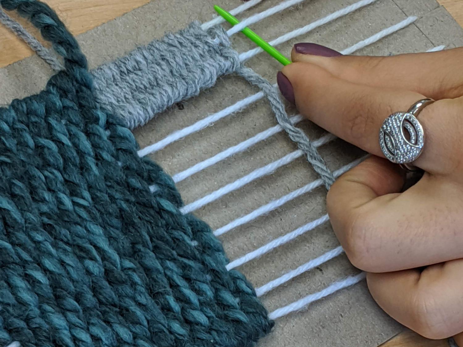A hand weaves yarn onto a small cardboard tapestry loom