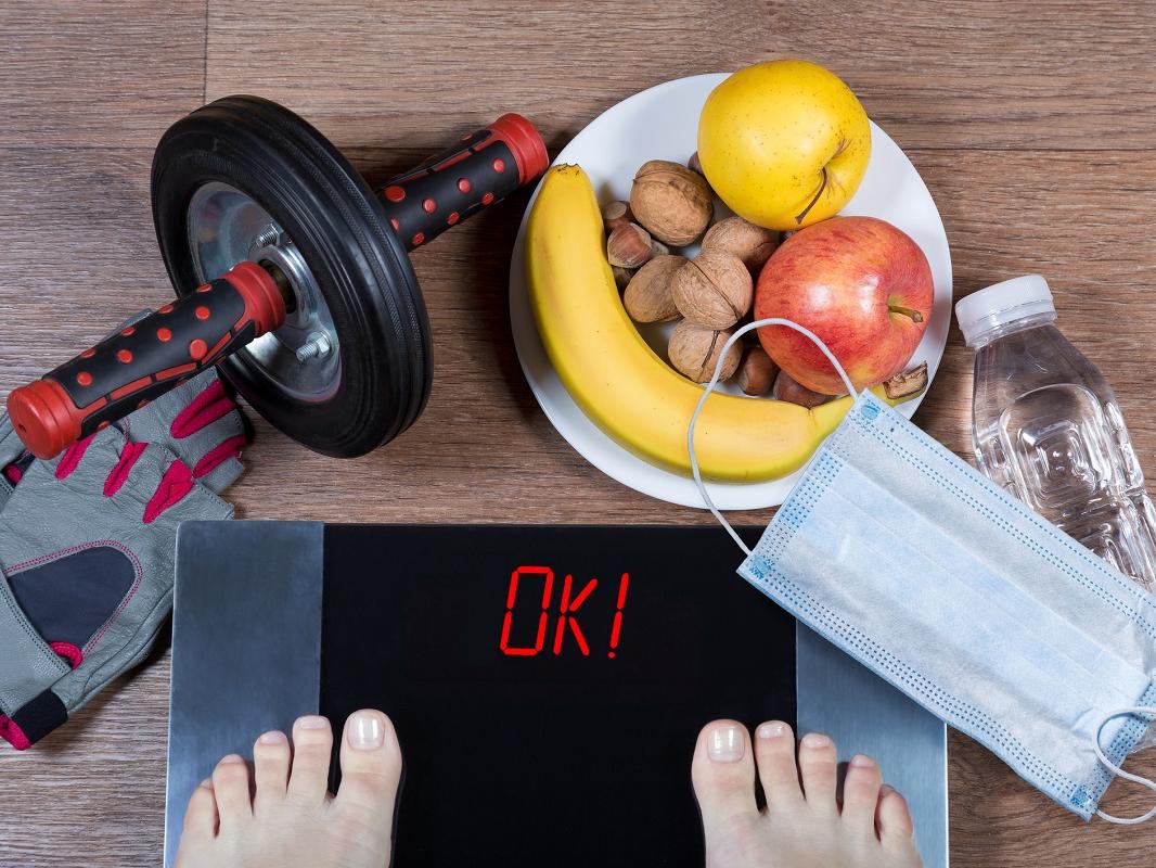 Person checks weight after quarantine. Digital scales with word ok surrounded by sport accessories, healthy food, water bottle and face mask.