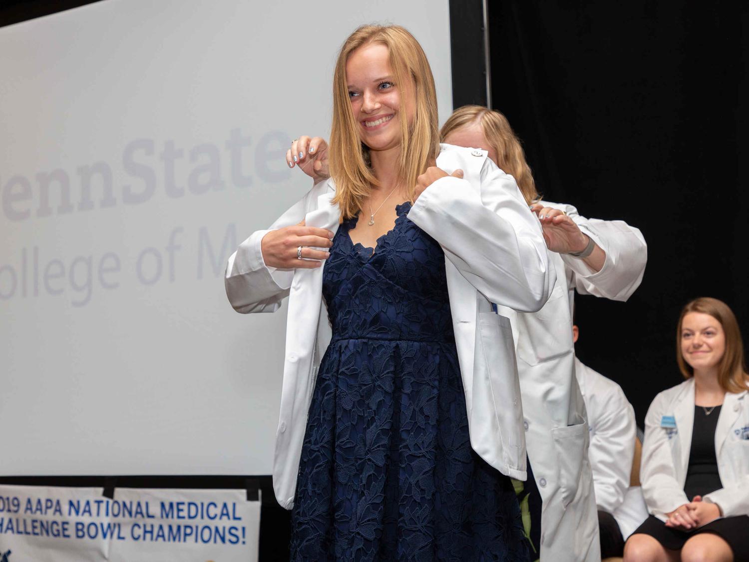 Kerri Schopf, wearing a dress, smiles as another woman stands behind her and slips a white coat onto her shoulders. Behind her, the words Penn State College of Medicine are projected onto a screen.