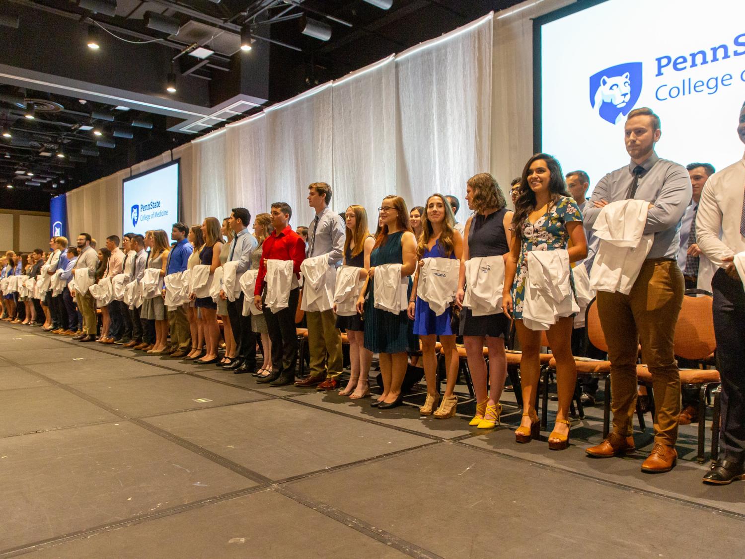 Several dozen medical students stand in two rows across a stage, with white medical coats draped over their left arms. The Penn State College of Medicine logo is projected on two screens behind them, and just in front of a pipe-and-drape backdrop.