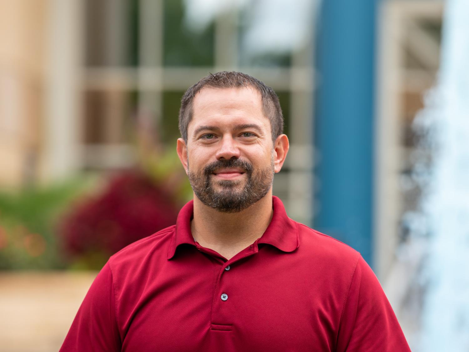 A headshot photo of Will Moyer II standing outside wearing a red shirt