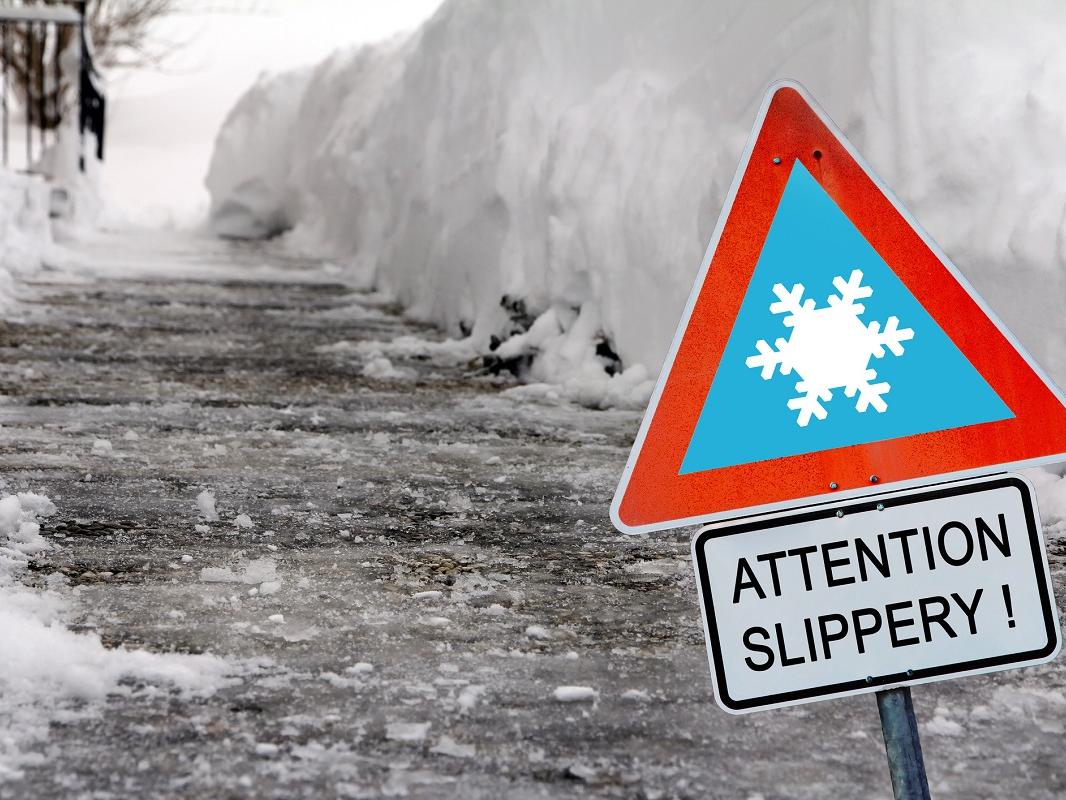 An icy sidewalk in winter with a sign in the foreground reading ‘Attention slippery!’