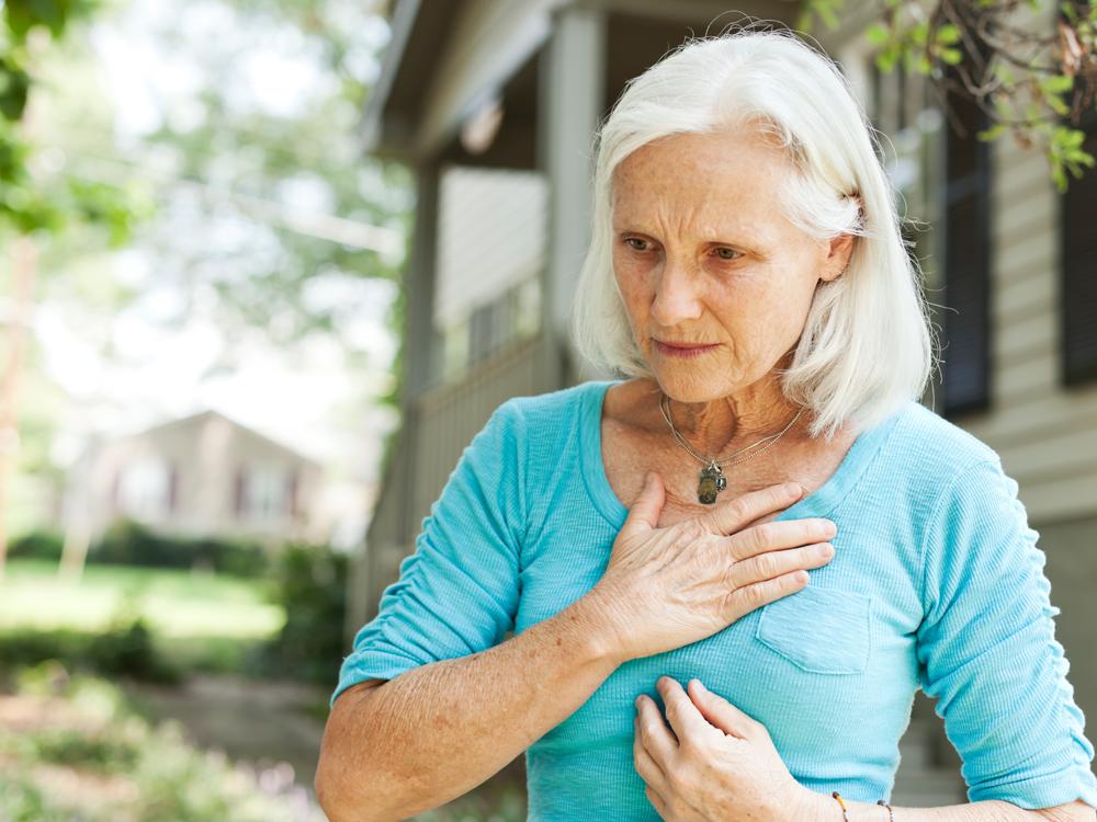A woman in a blue shirt stands outside of a home, which is slightly out of focus in the background. The woman holds her hand to her chest, up near her neck.