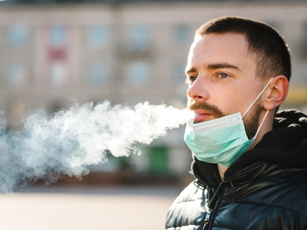 A bearded man with a paper mask below his mouth exhales smoke. 