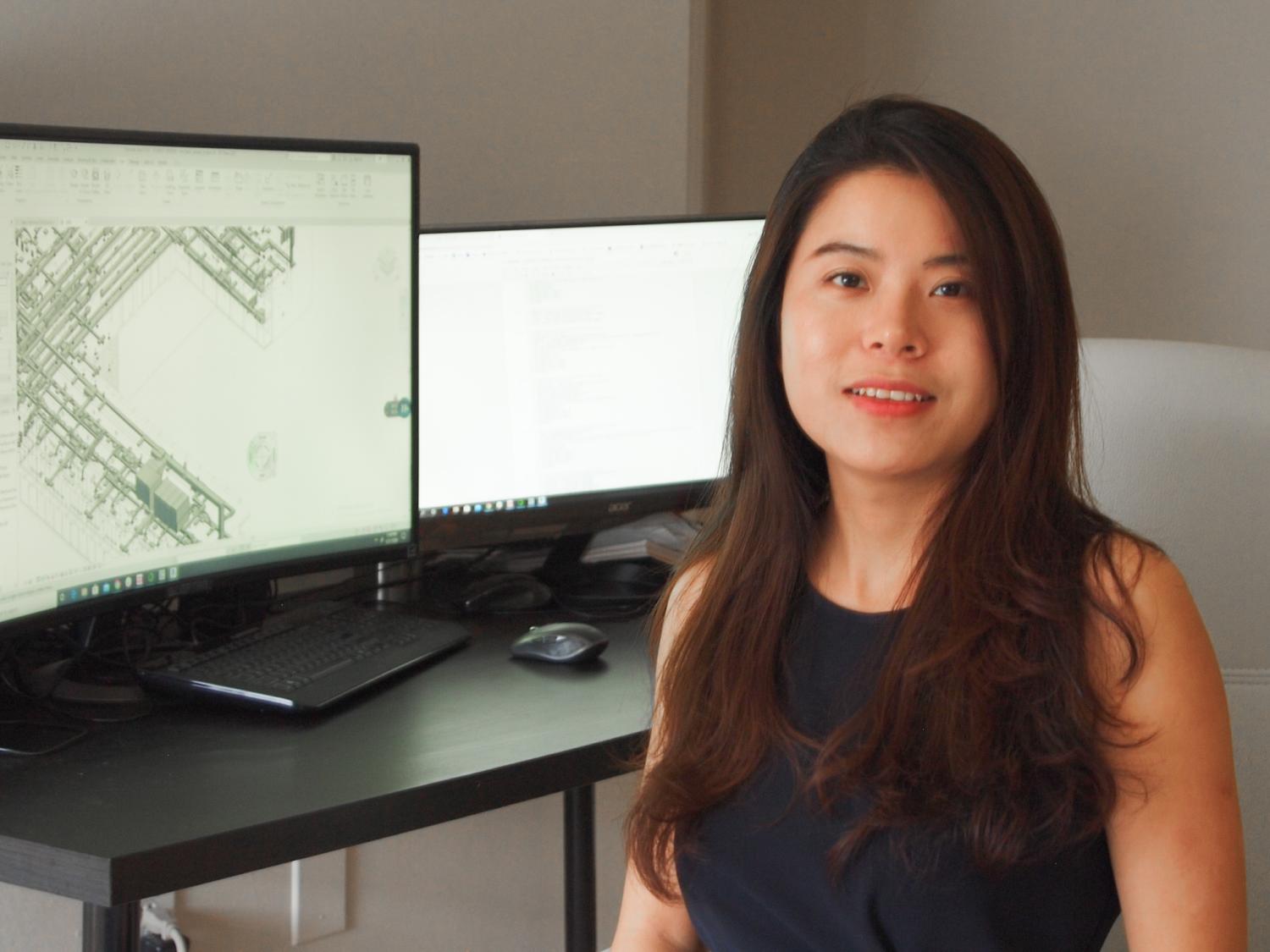 A young Asian woman sits at a desk with two computer monitors. 