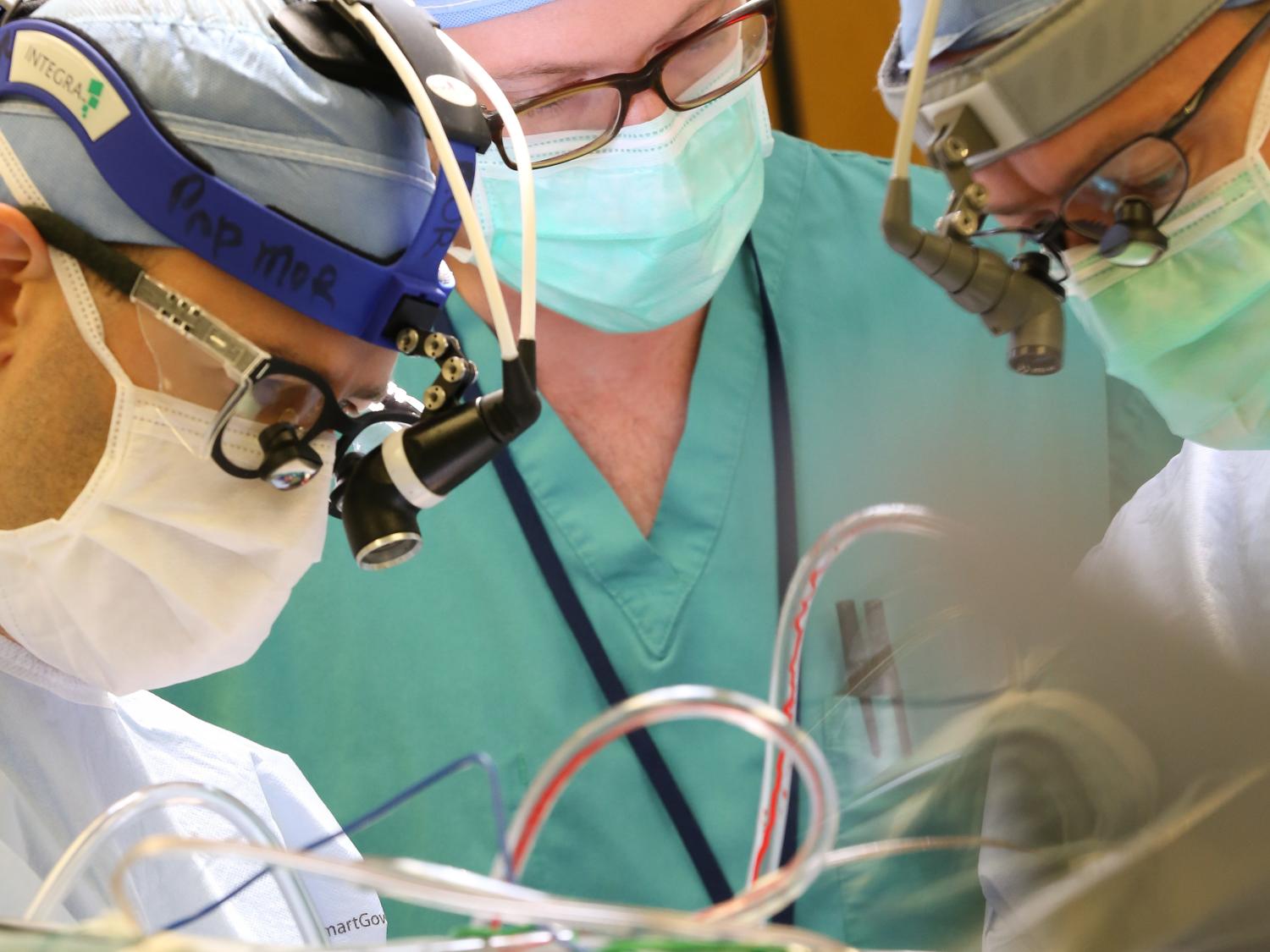 Three surgeons/medical staff work in the operating room, over a patient. The patient is not visible, but tubes emerge out of the bottom of the photo.