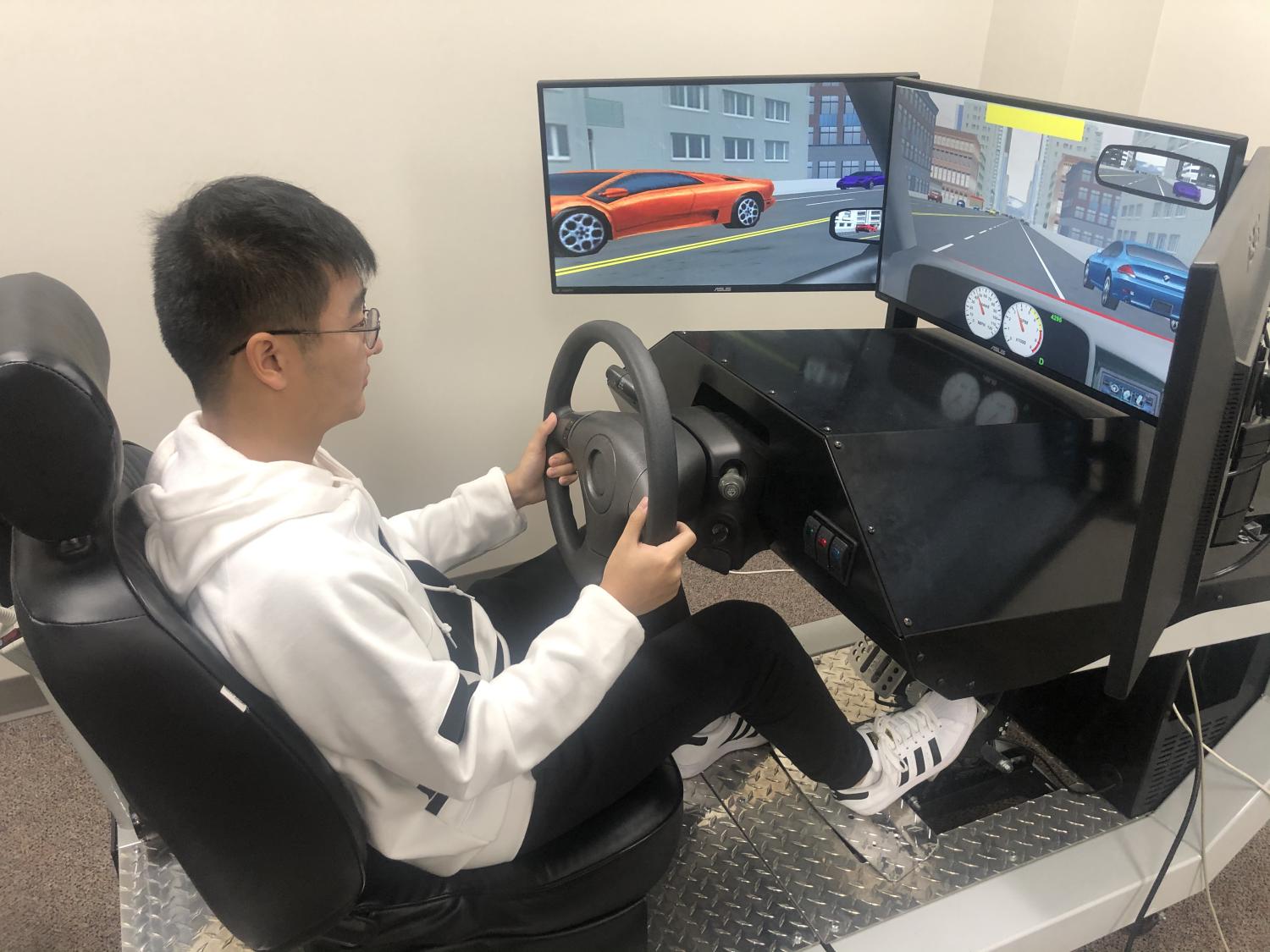 Man sits at test driving station with a steering wheel and computer screens to simulate driving on the road.
