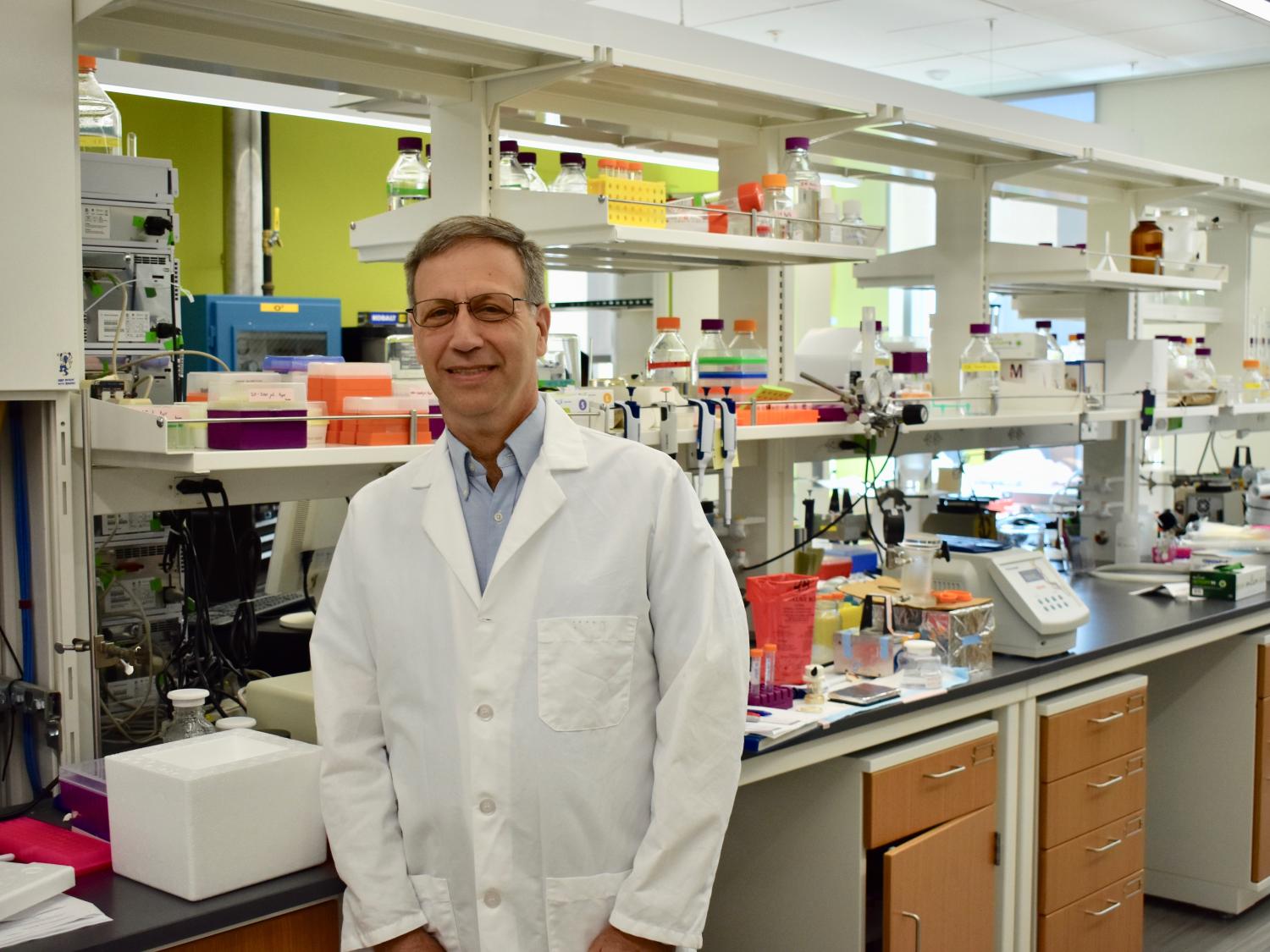 Man with glasses standing in white lab coat with lab equipment in background.