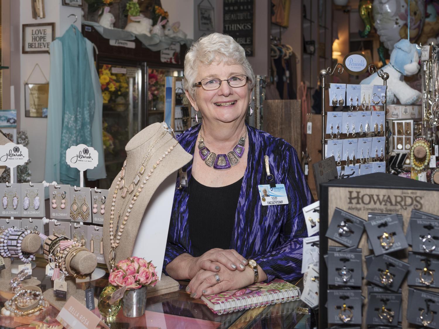 Sue Espenshade poses for a photo behind the counter at the Hershey Medical Center gift shop. Several racks of earrings and other jewelry are in the foreground; a case of flowers and various gifts in the background.