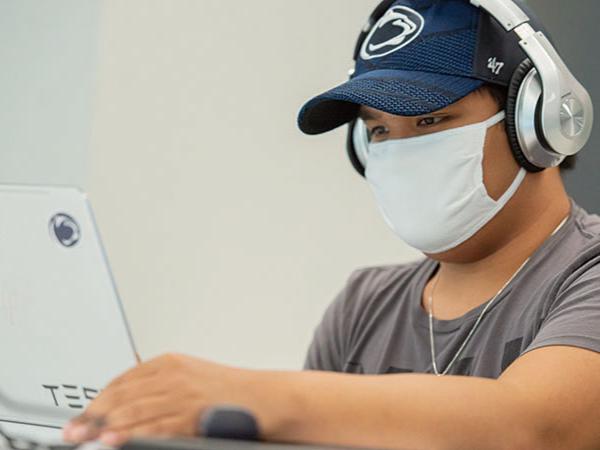 Male student in Penn State hat and headphones works on laptop in a student lounge. 