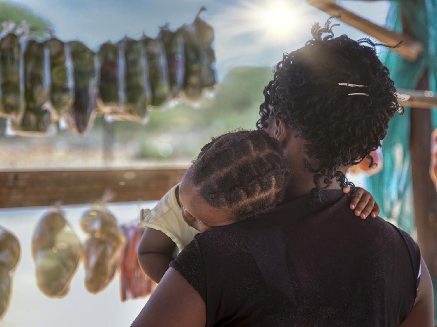 Woman seen from behind holding small child in her arms as she browses for food in an African market