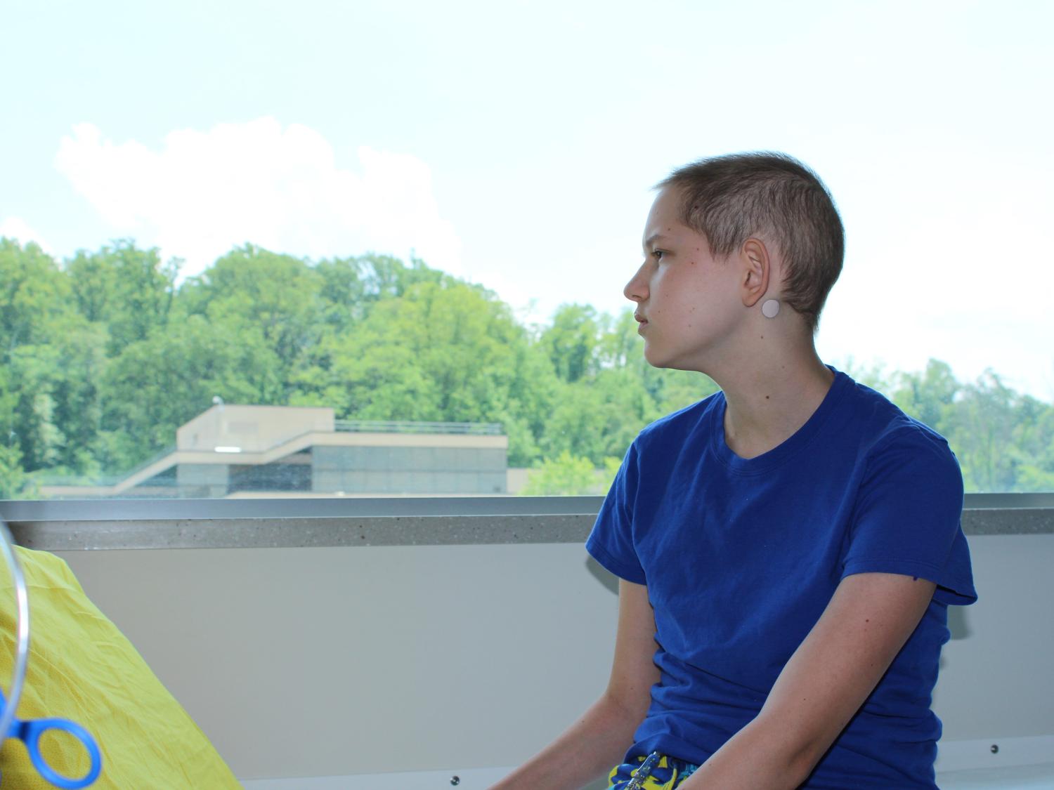 A teenage girl wears a blue t-shirt and sits sideways looking out of a large window.
