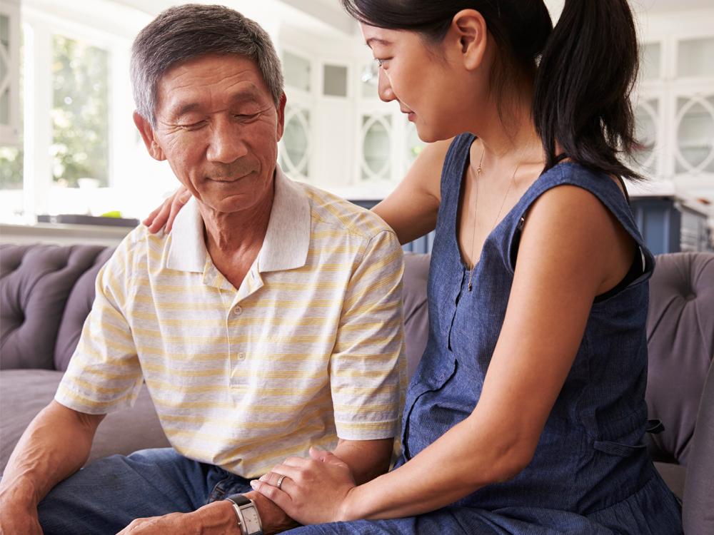 A young woman sits on a couch next to an older man. She has her right arm around him and looks at him. He has a neutral expression and looks downward.