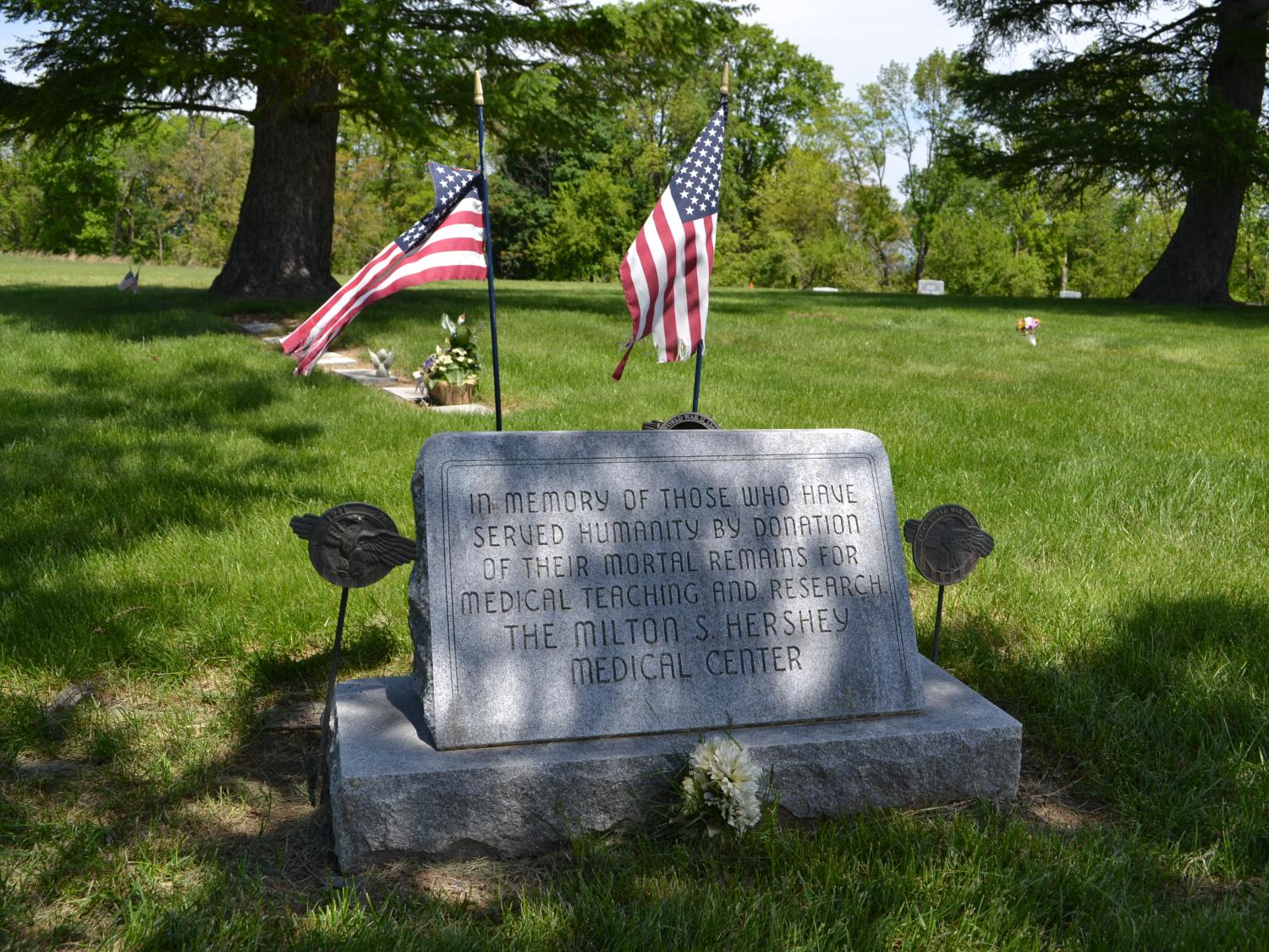 A stone is engraved with the words "In memory of those who have served humanity by donation of their mortal remains for medical teaching and research at the Milton S. Hershey Medical Center." Two American flags emerge from the top of the stone.