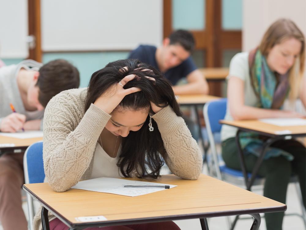 A teenage girl sits at a school desk, holding the top of her head with her hands, looking down at a paper on the desk. Three other students are in the background, slightly out of focus.