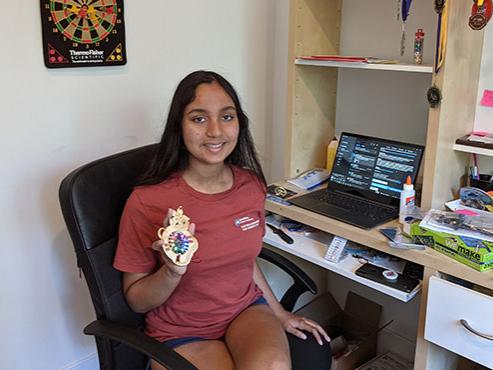 A girl in a maroon t-shirt sits in front of a laptop and holds up a small piece of wood with colorful lights. 