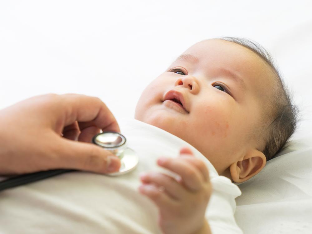 An infant wearing white lays on a white sheet. An adult is holding a stethoscope on the infant’s chest.