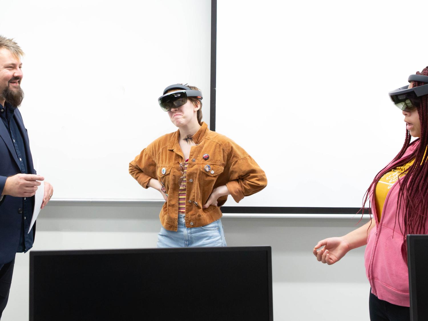 The professor stands to the left as two female students wear goggles as part of a classroom experiment.
