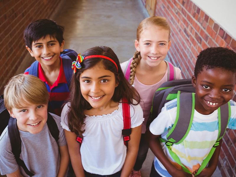 Five young children stand together in a school hallway, looking up and smiling at the camera.