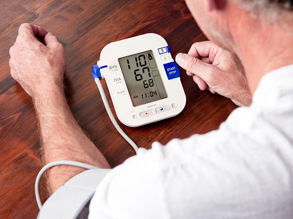A man sits at a table, looking down at the LCD display on a small blood pressure monitor. The cuff is wrapped around his upper left arm.