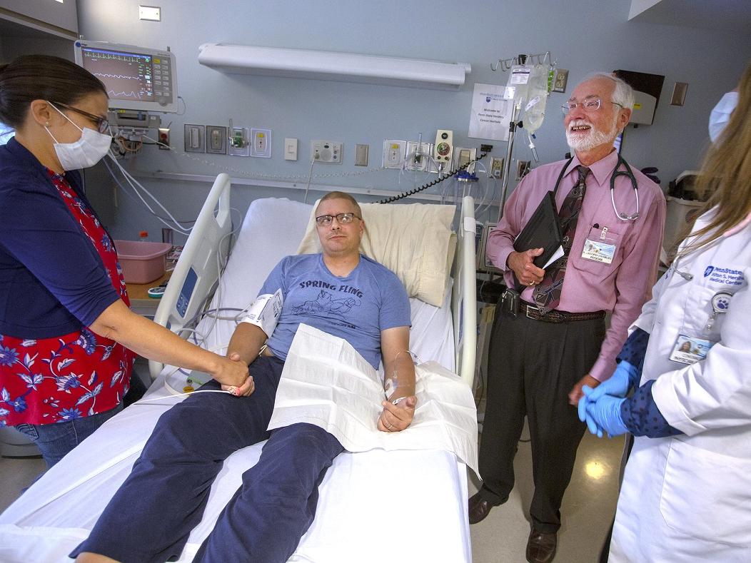 A wife holds her husband’s hand in a hospital room while talking with a doctor and physician assistant.