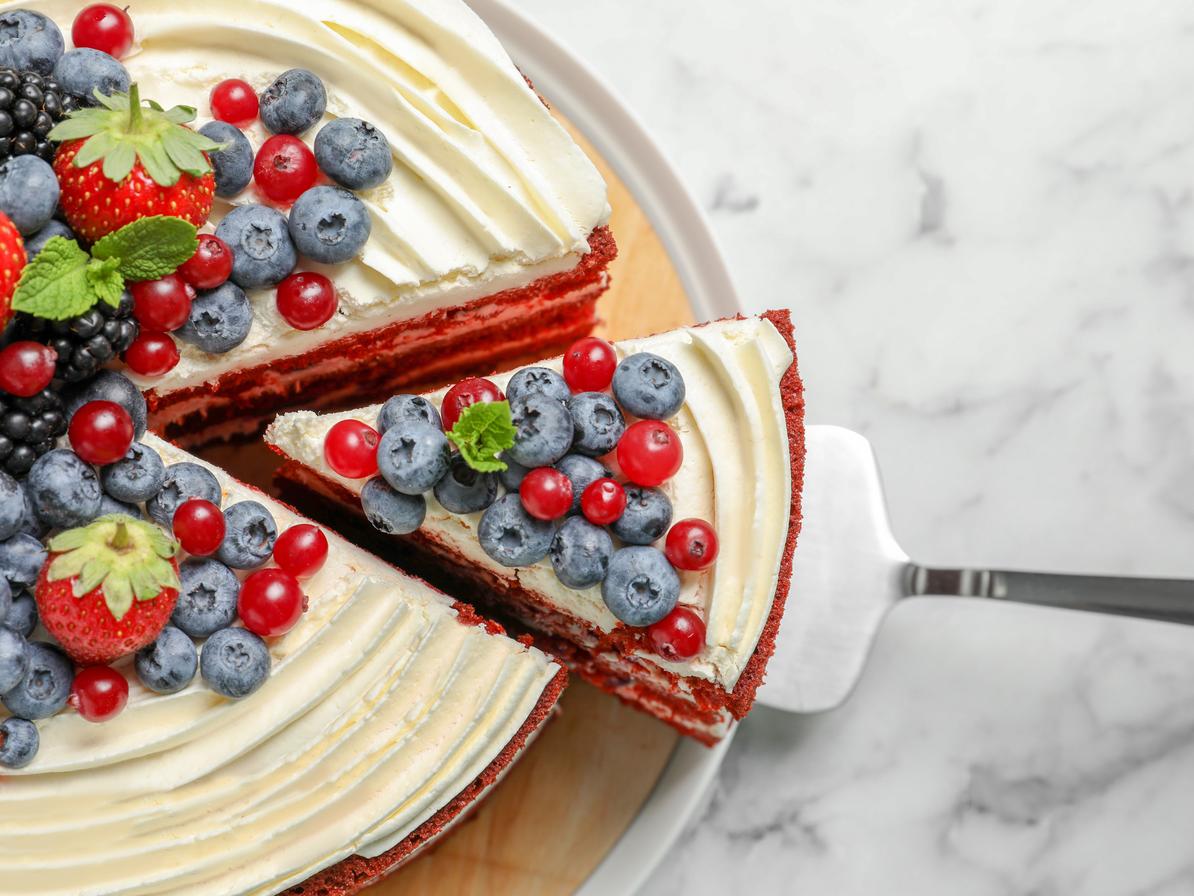 A triangular slice of a red velvet cake topped with berries if lifted from a platter with a silver cake cutter.