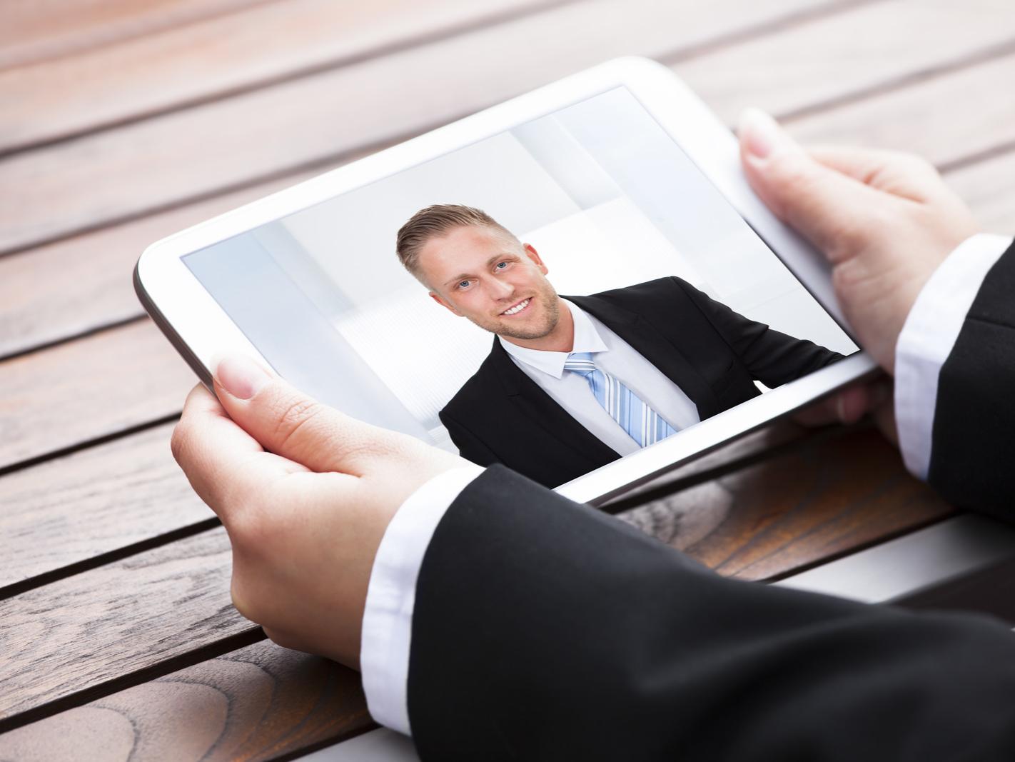 A man in a suit holds a tablet computer that shows another man in a suit.