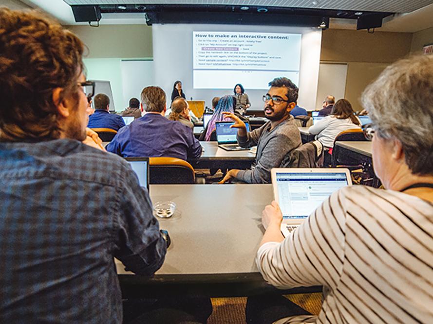 A group of people seated with laptops looking  at two presenters standing in the front of the room with an overhead presentation on the screen