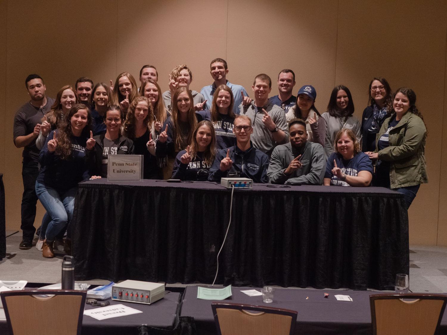 A group of approximately 25 people is pictured, with a few seated at a table and the rest standing behind them.