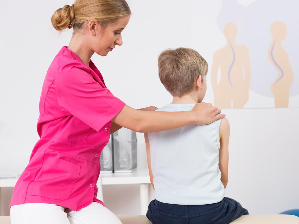 In a clinical exam room, a woman sits beside a young boy on an exam table. Her hands are on his shoulders and she appears to be looking at the boy’s spine.