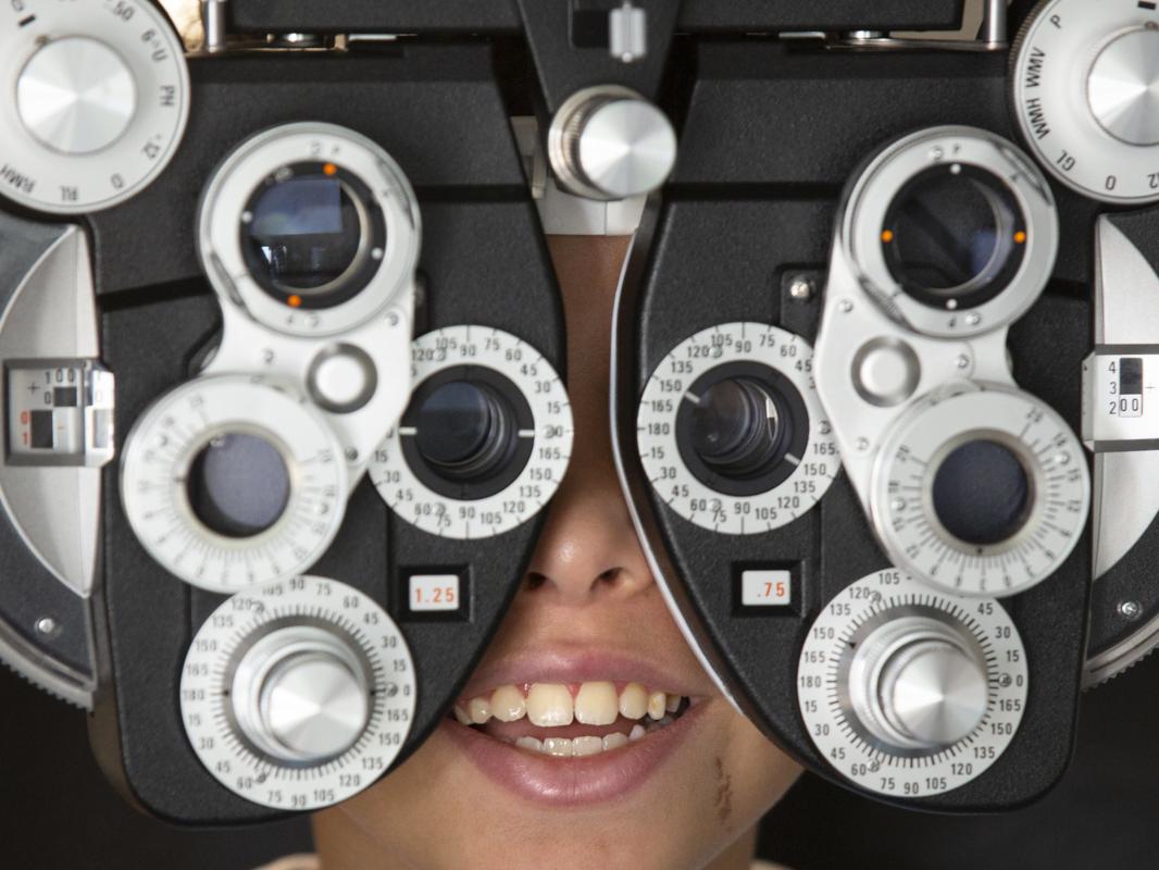 A close-up of a young child looking through the lenses of eye examination equipment, which contains several dials.