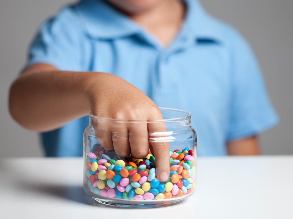 A jar of small, multicolored candies is in the foreground. A young boy stands behind the jar with his hand extending into it.