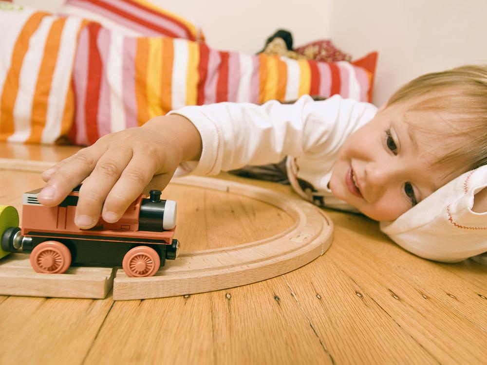 A young child lays on his side on a wooden floor, with his hand on a train set, which is in the foreground. A bed with a multi-colored, striped bedspread is in the background, slightly out of focus.