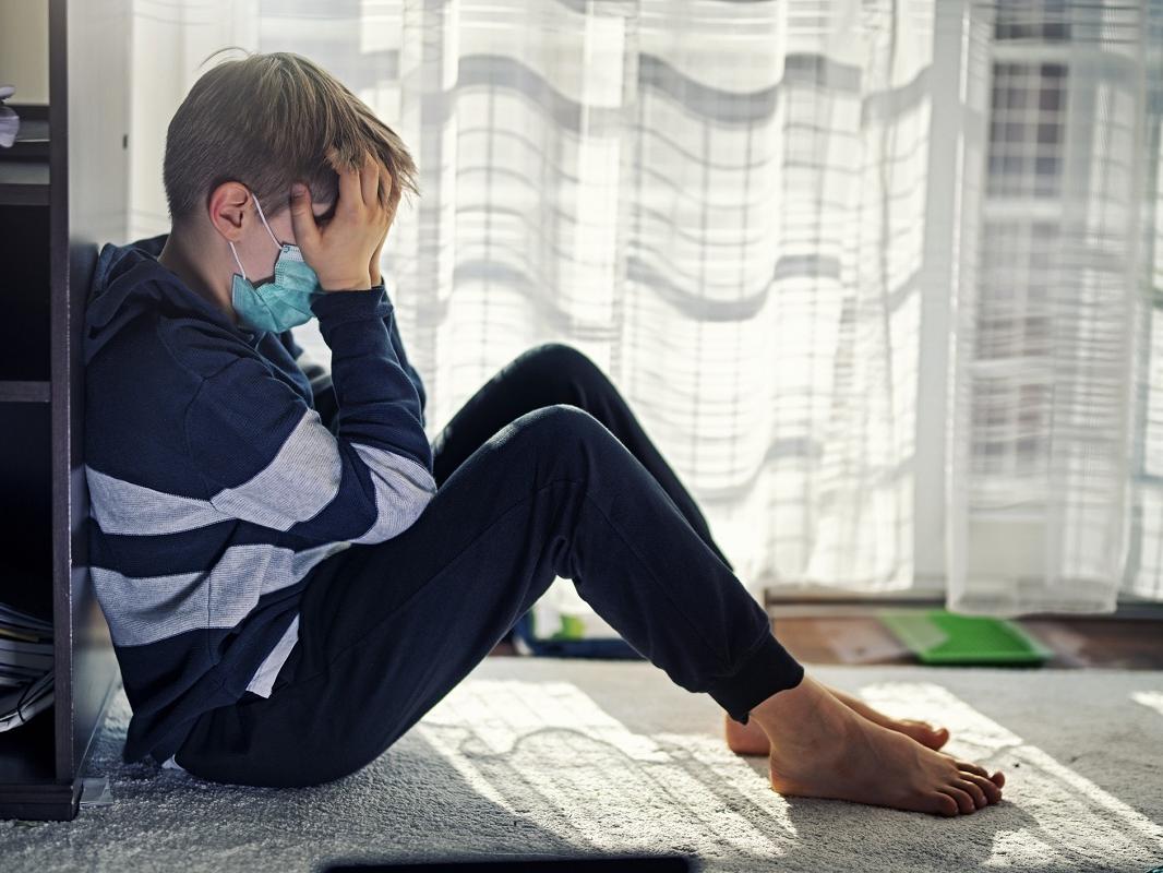 A young boy sits by a window, wearing a surgical mask, with his face in his hands.