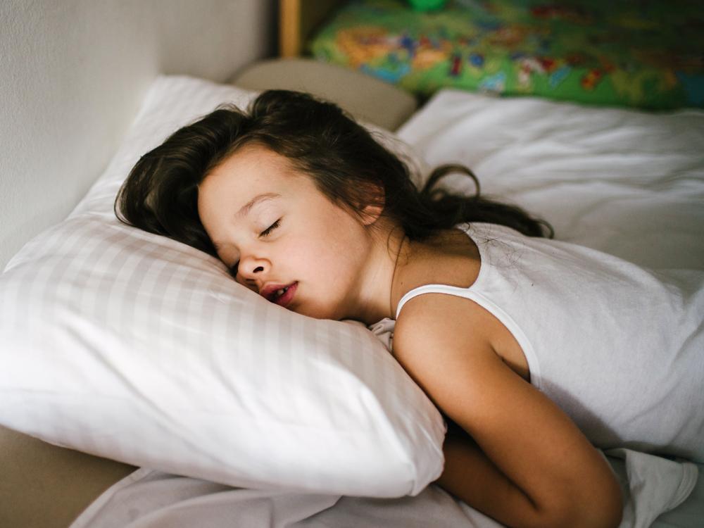 A young girl sleeps in bed, with her head on a pillow. Her head is facing the camera.