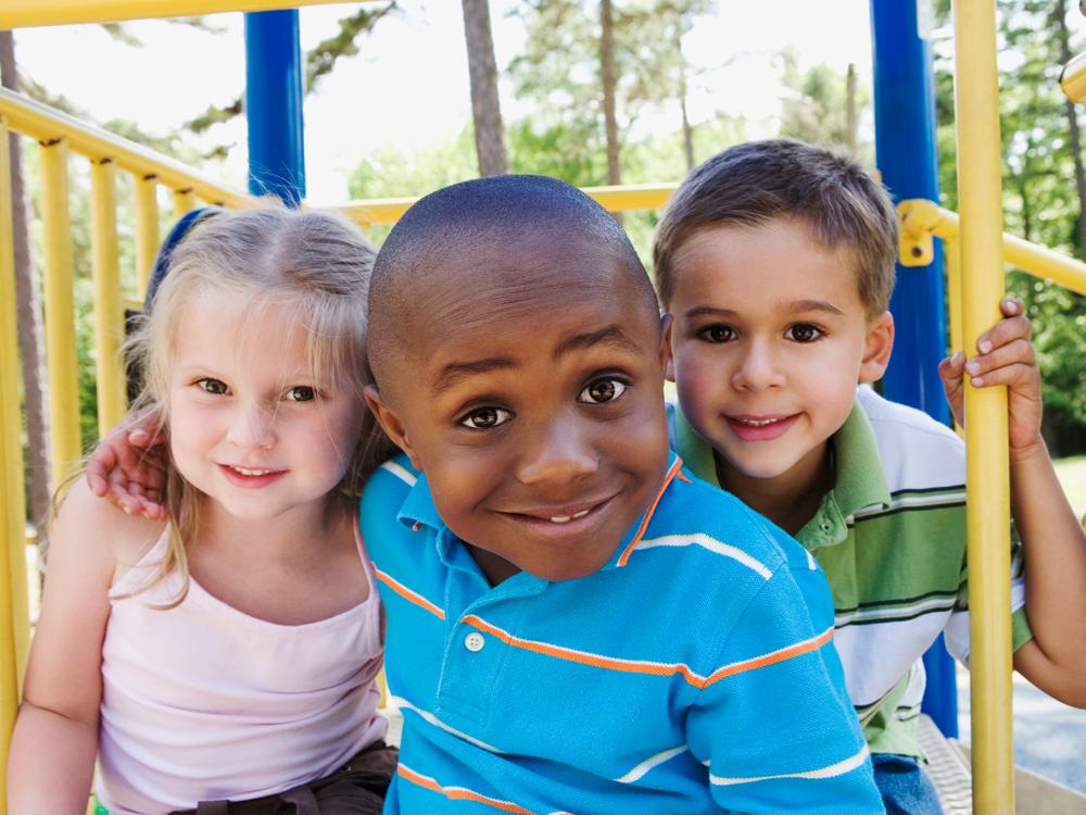 Three young children pose for a photo while sitting on a blue and yellow piece of playground equipment. A wooded area and a sunny sky are in the background.